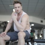 Serious young muscular sportsman in sportswear resting on bench during intense weightlifting workout in modern gym and looking at camera